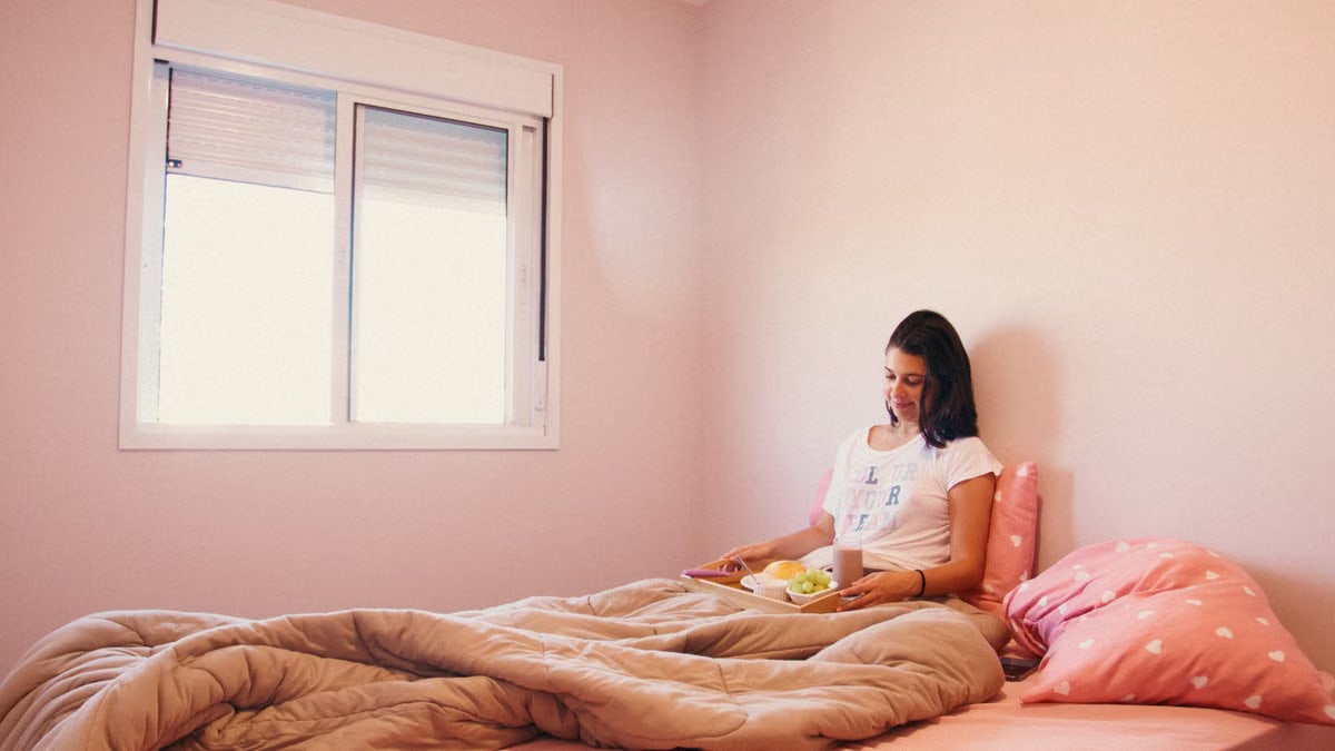 A woman relaxes with breakfast in bed in a cozy pink room, enjoying a peaceful morning.